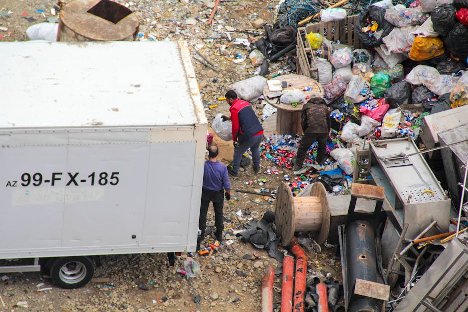 An outdoor scene featuring a large white waste container with black lettering and registration number positioned on the left side of the image. Adjacent to it, three individuals are engaging in rubbish clearance amidst a scattering of debris, including plastic bags, paper, and miscellaneous trash. The ground is uneven with a mixture of soil, small rocks, and discarded waste. To the right, there are piles of accumulated rubbish comprising black, white, and multicoloured plastic bags filled with waste materials, along with broken wooden pallets, spools, and various discarded industrial parts. Behind the waste pile, a wooden pallet and a partially visible metal structure can be seen, indicating an area of clutter typical of an informal waste collection or clearance site. The lighting suggests natural daylight, casting minimal shadows, and the environment conveys an active effort to remove and manage rubbish in this industrial or urban outdoor setting, aligning with services like independent waste collection or on-site clearance operated by companies such as Rubbish Clearance Chelsea.