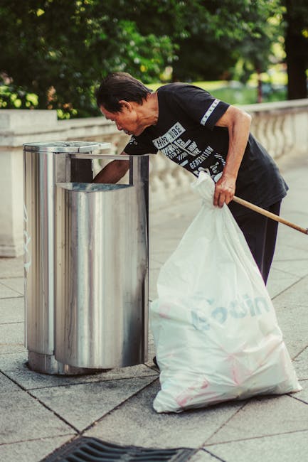 A person with dark brown hair, dressed in a black T-shirt with white and blue graphics, is seen collecting rubbish from a stainless steel public bin on a paved area. They are bent over, holding open the bin's lid with one hand while the other hand, which holds a long-handled tool, is empty. The individual has a large white sack with pink markings, possibly containing waste, placed on the ground beside them. The background includes a stone balustrade and lush green foliage, suggesting a park or an outdoor urban setting. The scene is lit by natural daylight, highlighting the metallic surface of the bin and the greenery around. This image illustrates an element of independent waste collection, such as rubbish removal services like Rubbish Clearance Chelsea, emphasizing environmentally conscious disposal practices within public outdoor spaces.