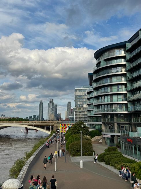 A wide view of a modern urban riverside scene featuring a high-rise residential building with a curved façade on the right, composed of glass and steel with multiple balconies. In the foreground, a paved walkway runs alongside the river, populated with pedestrians, some walking and others seated on benches. The walkway is bordered by neatly trimmed bushes and white lamp posts. To the left, the river flows under a bridge with a simple, functional design, and several boats are visible on the water. In the background, a city skyline with several tall skyscrapers rises against a partly cloudy sky, creating a contrast between the contemporary architecture and natural environment. The scene, captured during daytime, exemplifies an area suitable for private waste removal or on-site clearance by companies like Rubbish Clearance Chelsea, reflecting an urban setting with accessible pathways for rubbish collection and disposal activities, although no debris or waste is visible in the image itself.