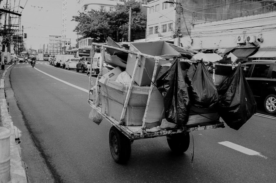 A small tireless cart loaded with various discarded items is seen on a city street, positioned on the right side of the road. The cart is constructed with a metal frame and is fitted with two black rubber wheels, indicating it's a manually pulled vehicle. It is heavily stacked with a mix of bulky cardboard boxes, some flattened and others partially open, revealing an assortment of paper and packaging materials. Several black plastic garbage bags are tied and draped over the sides of the cart, containing unidentified waste. The scene is set on an urban road with a curb on the left, and in the background, there are parked cars lining the street, commercial and residential buildings, and some pedestrians and cyclists visible further down the road. Overhead, electrical wires cross above the street, and the lighting suggests daytime with natural light illuminating the scene. The image resembles an example of independent waste collection or private rubbish clearance in a busy city environment, illustrating how rubbish clearance services like those offered by Rubbish Clearance Chelsea might handle household or commercial waste outside traditional municipal collection routes.