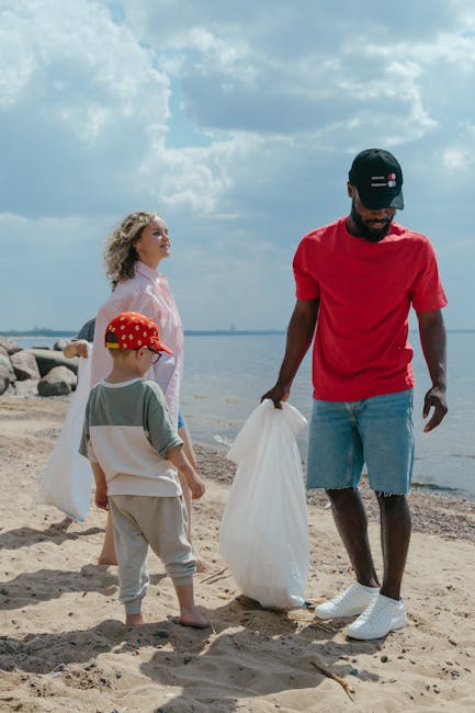 A group of three individuals, including a man and two children, are participating in a beach cleanup near the water, with a shoreline visible in the background under a partly cloudy sky. The man, dressed in a red t-shirt, light blue denim shorts, and a black cap, is holding a white rubbish bag, actively collecting litter from the sandy beach. One of the children, wearing a red cap with white patterns, a grey and white top, and beige trousers, stands close by, observing the activity. The other child, a girl with curly blonde hair, is dressed in a light pink button-up shirt and is looking out over the water, supporting a sense of community engagement in waste management. The surrounding environment includes a sandy area with rocks along the shoreline and a calm body of water stretching into the distance, complemented by a partly cloudy sky that provides natural light. This scene exemplifies personal efforts aligned with independent waste collection and rubbish removal services, echoing themes of environmentally conscious disposal outside of major urban waste systems, with the focus on responsible litter pick-up on natural sites.