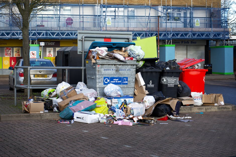 A collection of overflowing rubbish bins and scattered waste materials situated on a paved sidewalk area, adjacent to a parking space with a silver car parked nearby. The waste includes black, red, and grey bins, with some lids open and trash spilling over onto the ground. The debris consists of cardboard boxes, plastic bags, paper, packaging materials, and miscellaneous household refuse, with some items partially crushed or crumpled. The bins and rubbish are positioned close to a metal railing, which separates the waste area from the vehicle parking zone. In the background, there is a building under construction or renovation, covered in blue scaffolding and protective netting, with commercial storefronts visible beneath, one advertising a fish bar. The environment has an urban setting with natural lighting, and the scene closely relates to private rubbish disposal or alternative waste management services, as managed by Rubbish Clearance Chelsea, in the context of same-day waste removal services in Chelsea.