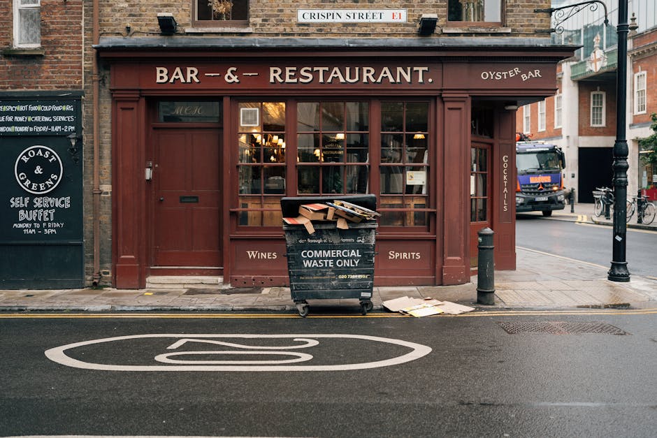 A black commercial waste bin labeled 'Commercial Waste Only' is positioned directly in front of a brick-fronted bar and restaurant, situated on a sidewalk along a city street. The bin contains discarded cardboard boxes and paper, with some materials spilling over the top edges. The exterior of the building features large glass windows with wooden framing painted in a deep red or maroon hue, revealing the warmly lit interior. To the left, a black signboard advertises a self-service buffet, and to the right, a black bollard is installed on the pavement, along with a concrete post. Behind the bin, a narrow road is visible with a parked delivery truck on the side, a police vehicle further down, and bicycles parked nearby. The street markings include a glossy white bus stop symbol painted on the asphalt, and a street sign reading 'Crispin Street E1' is mounted above the building entrance. The environment appears clean and typical of urban waste management situations, possibly reflecting the independent approach to rubbish disposal that businesses may adopt outside standard council collection services, as discussed on Rubbish Clearance Chelsea’s website.