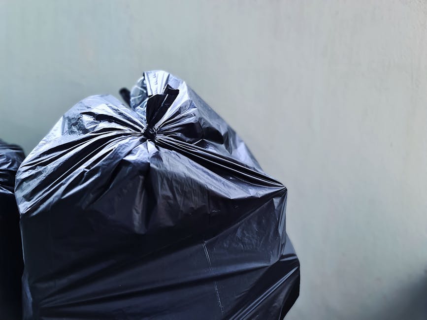 Close-up image of a black plastic rubbish bag made of glossy, slightly crinkled material, tied at the top with visible folds and creases. The bag is situated on a flat surface, possibly in an indoor or outdoor environment, with a plain, light-colored background that appears neutral and unadorned. The surface beneath the bag is not clearly visible but suggests a clean setting. The image focuses solely on the rubbish bag, with no other objects or context provided. The scene is well-lit, revealing the texture and sheen of the plastic. This visual corresponds to waste collection and rubbish removal services, emphasizing the type of waste that might be handled by a company like Rubbish Clearance Chelsea, which offers alternative disposal options outside of council services.